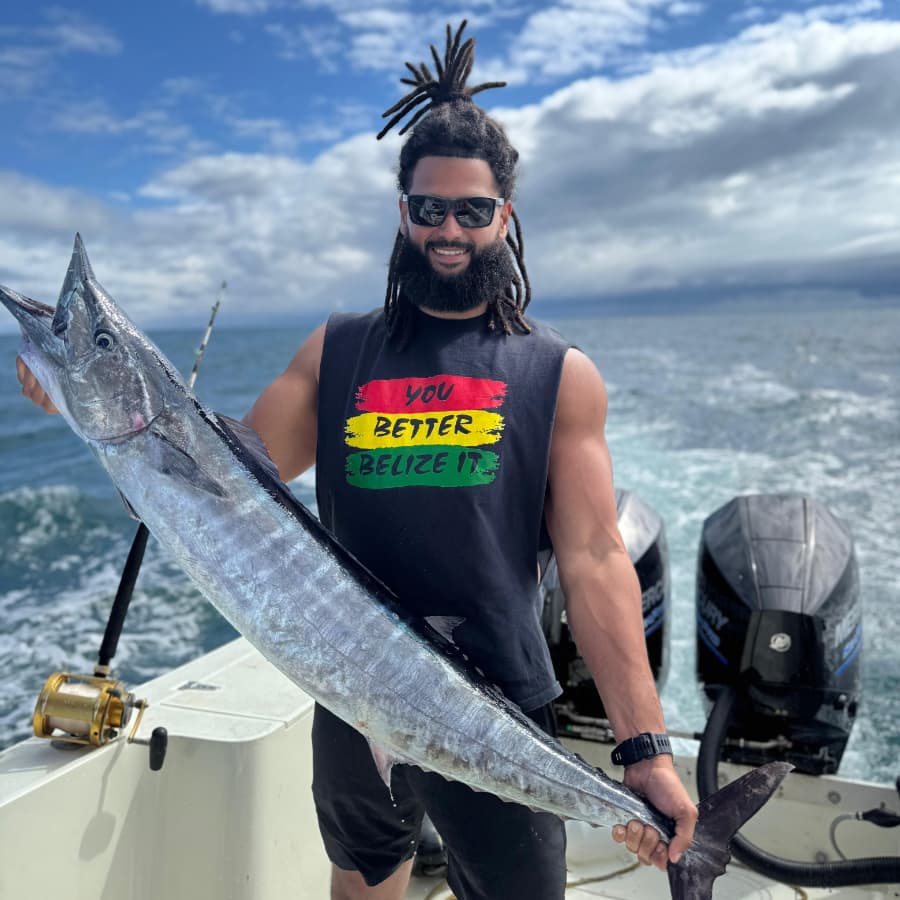 A man with dreadlocks holds a large fish on a boat, wearing sunglasses and a tank top.