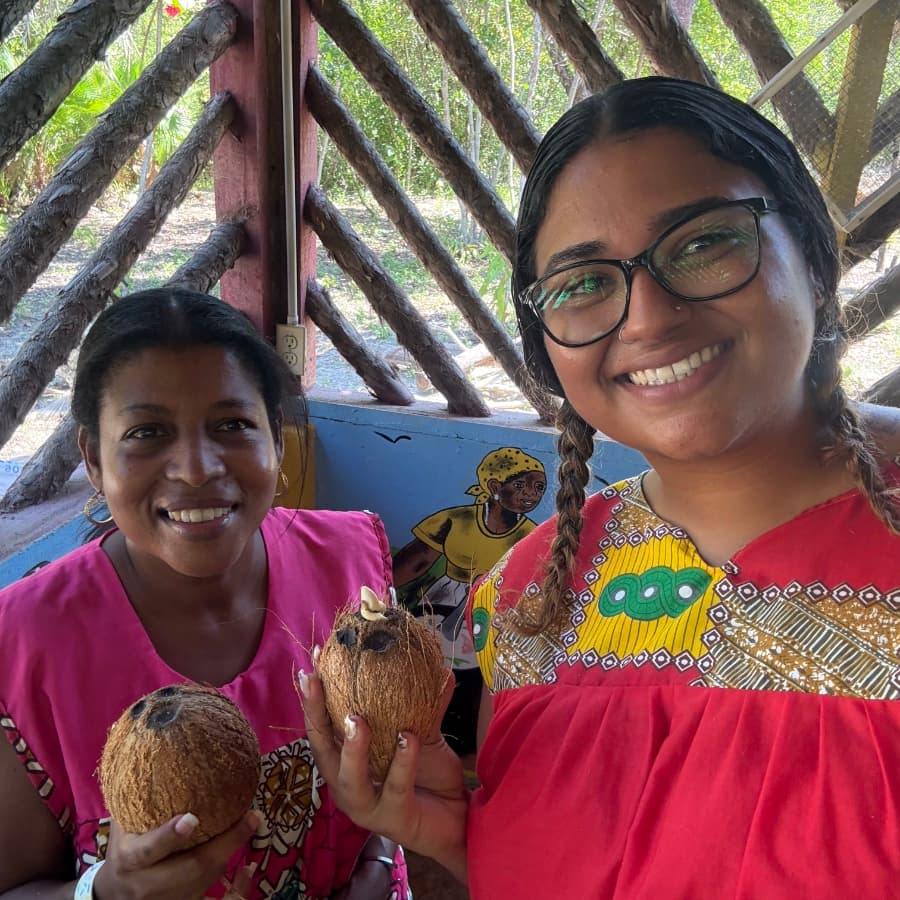 Two women smile while holding coconuts in a colorful, rustic setting.