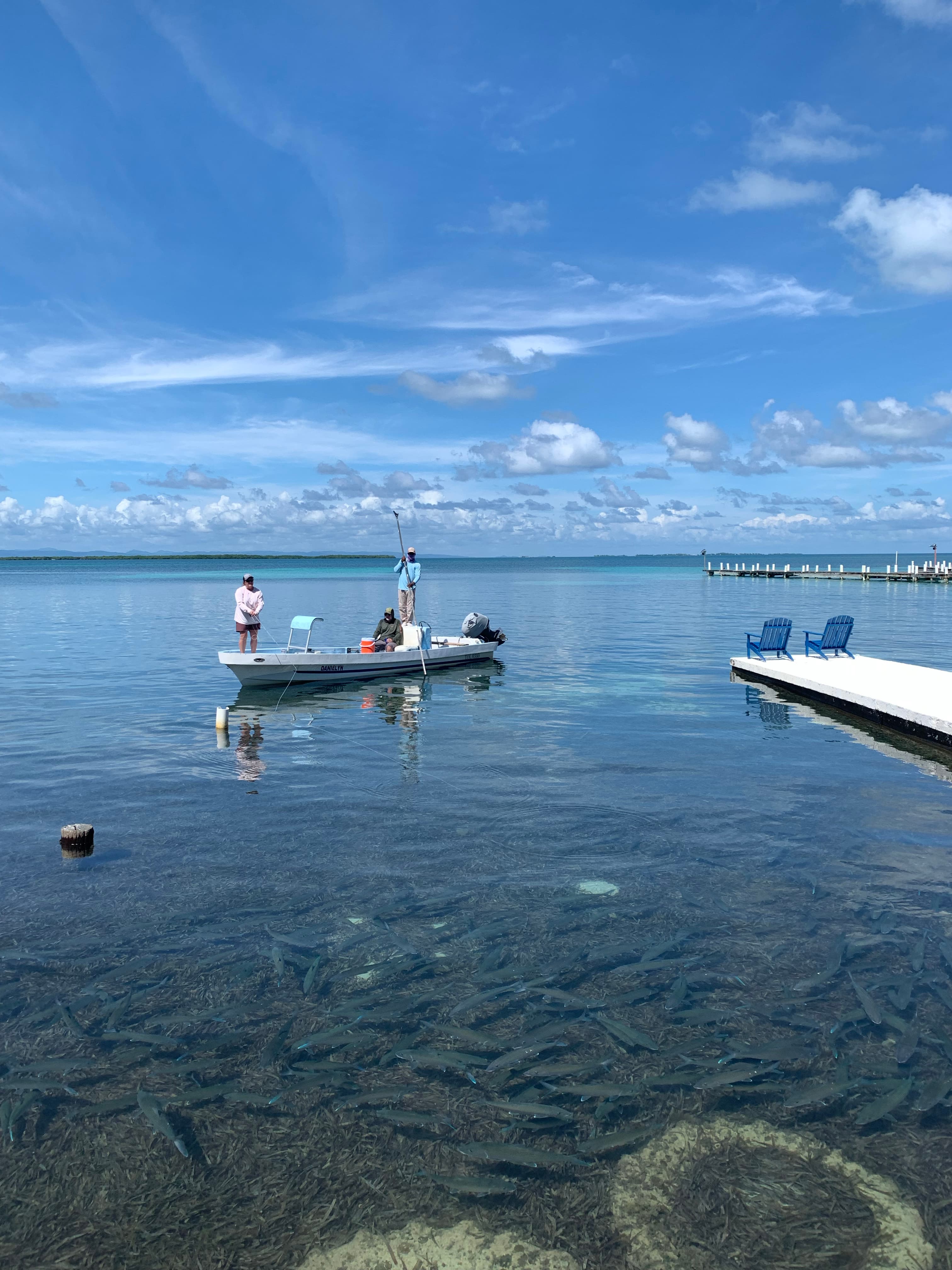 A boat is anchored in clear water with two people fishing, surrounded by schools of fish beneath the surface.