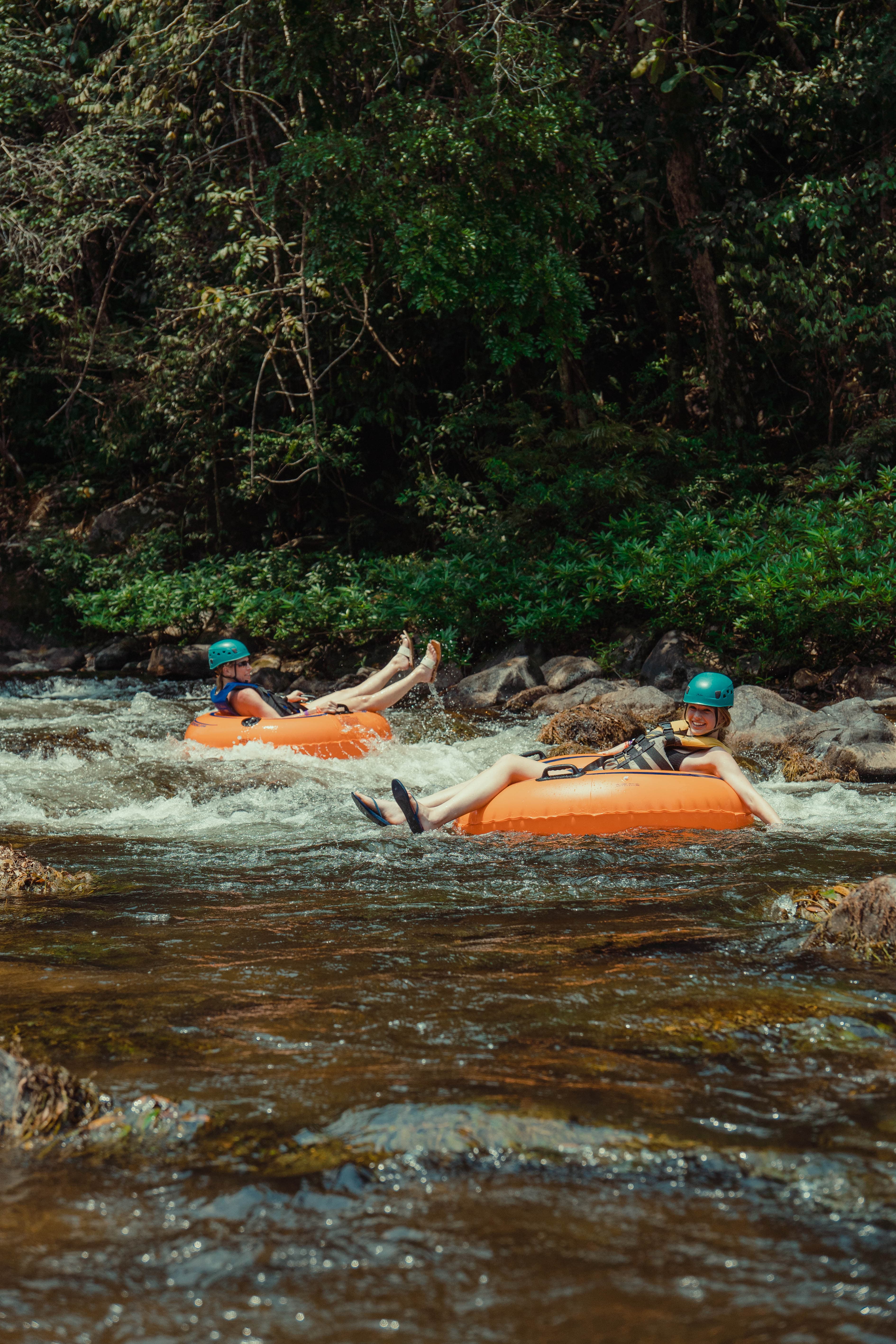 Two people float on orange inner tubes down a river surrounded by lush greenery.