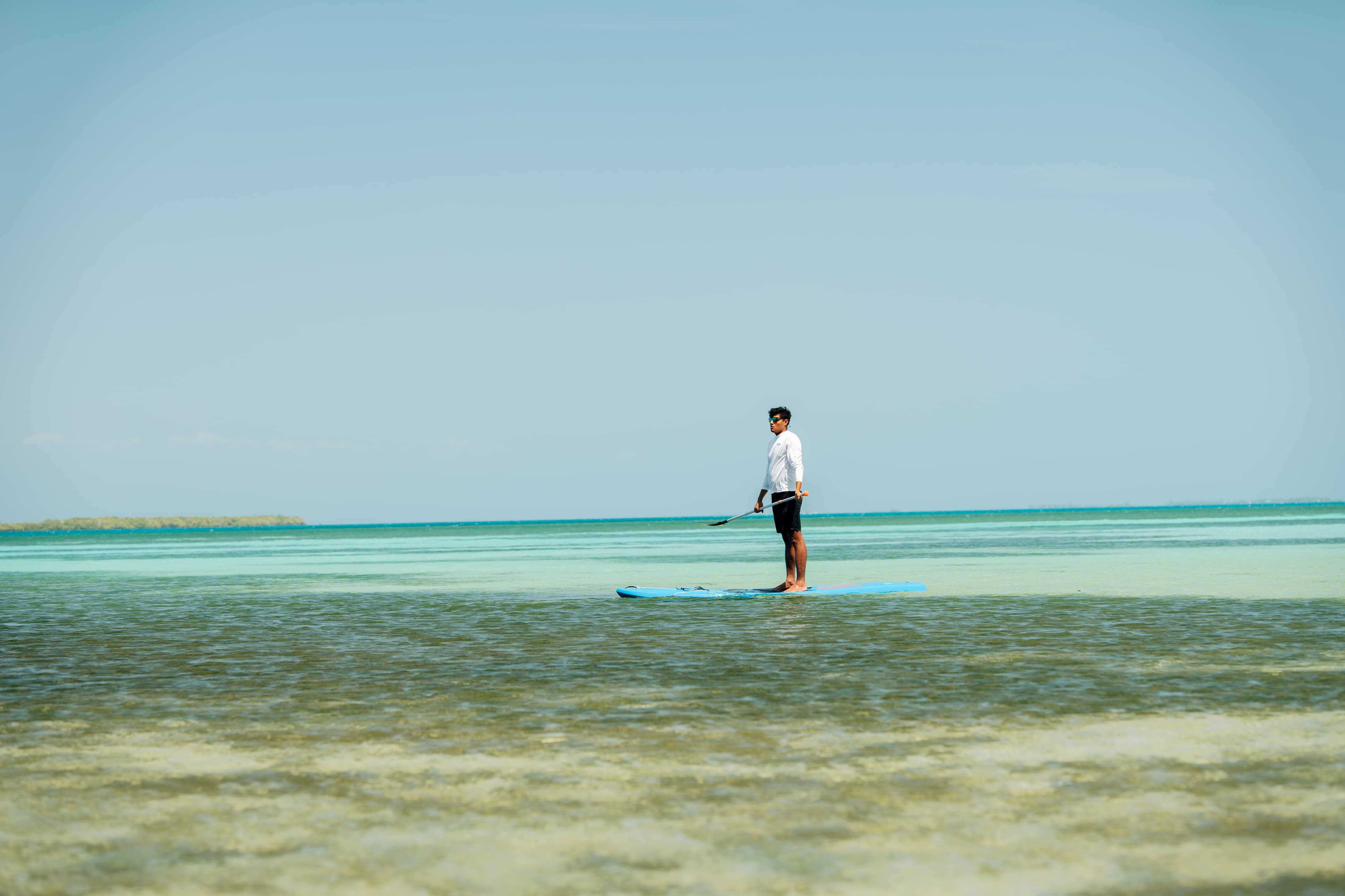 A person stands on a paddleboard in shallow water, surrounded by turquoise and green hues of the ocean.