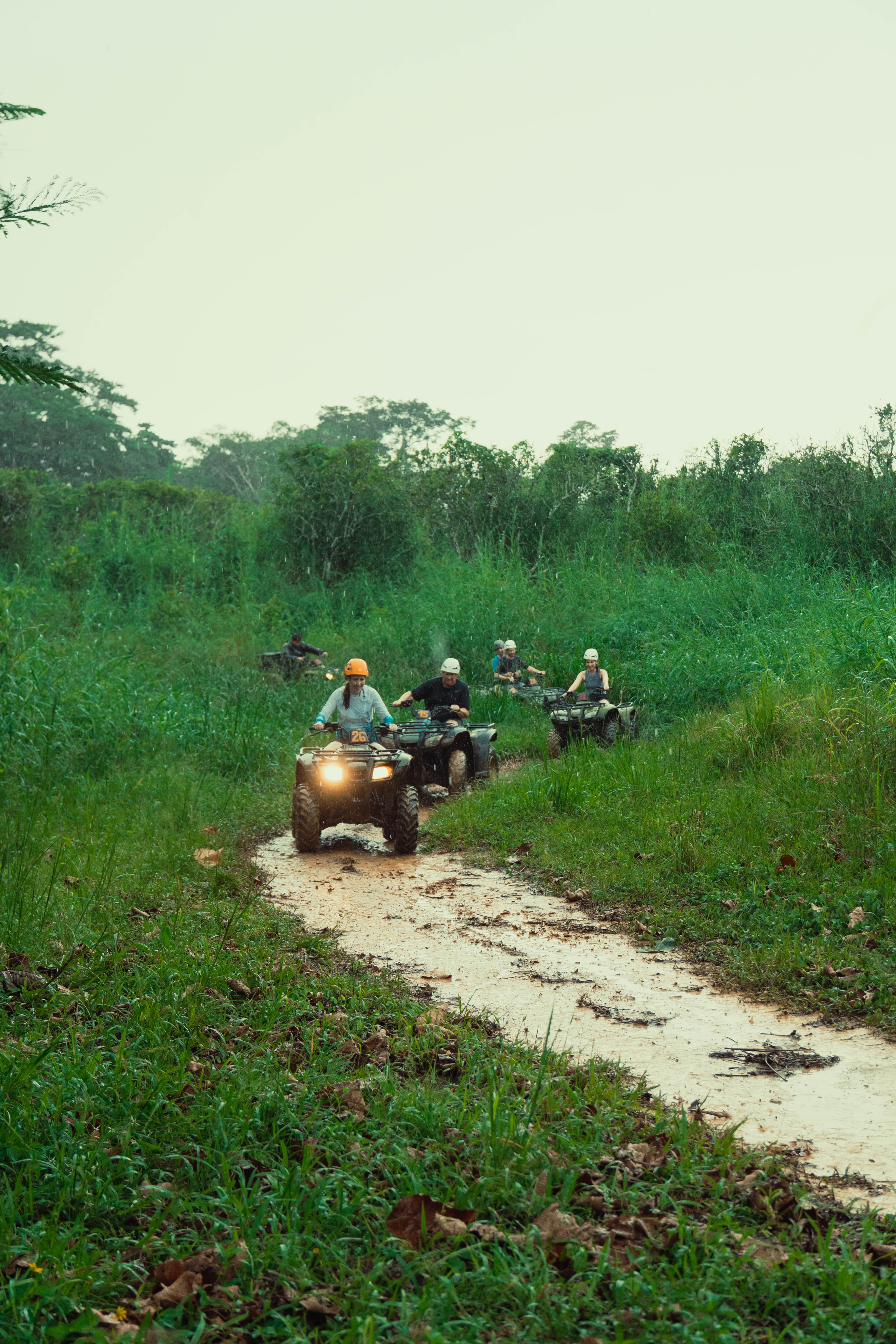 A group of people rides ATVs on a muddy path surrounded by lush green vegetation.