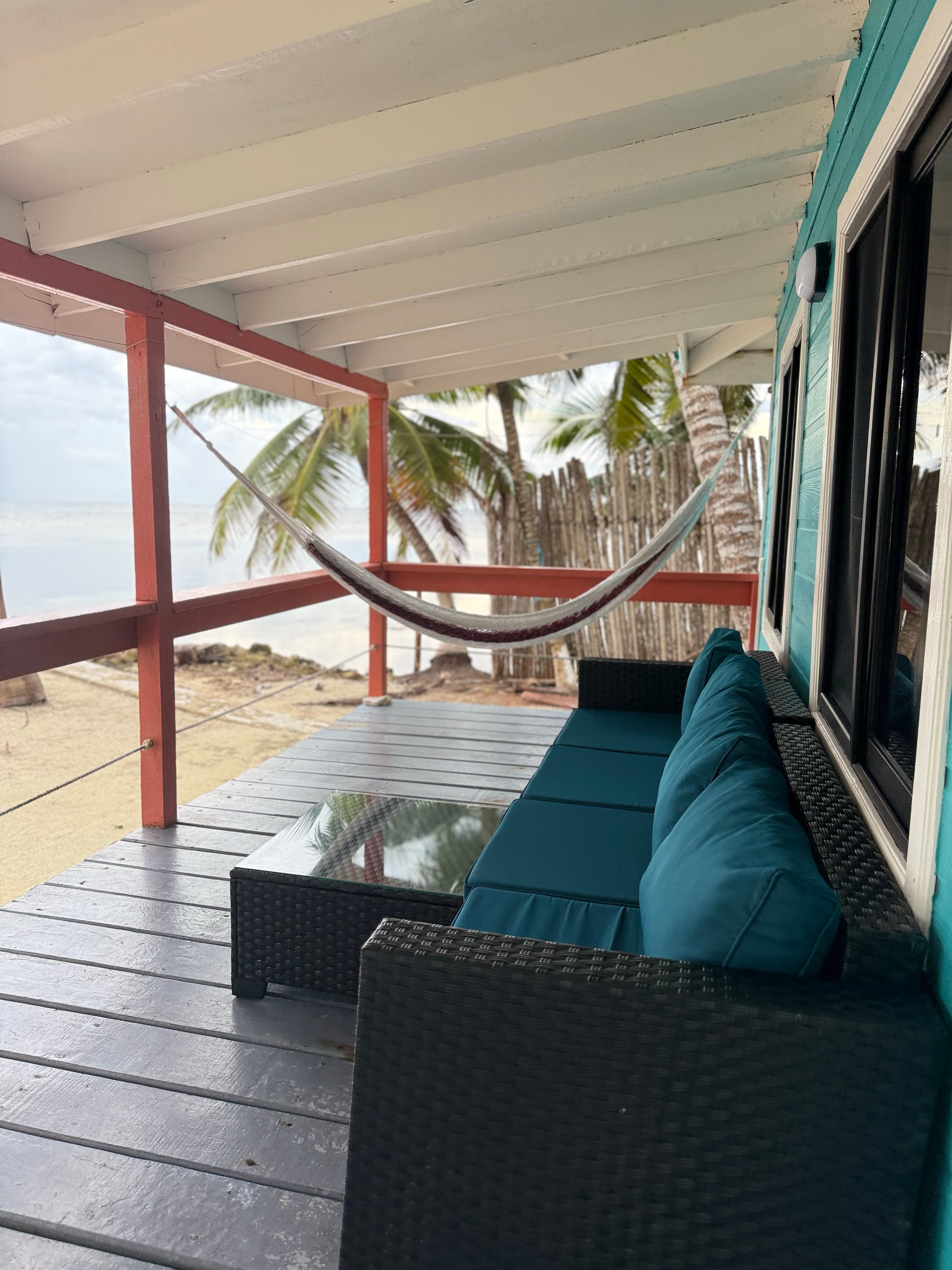 A cozy porch with a couch and glass table overlooks a beach and palm trees.