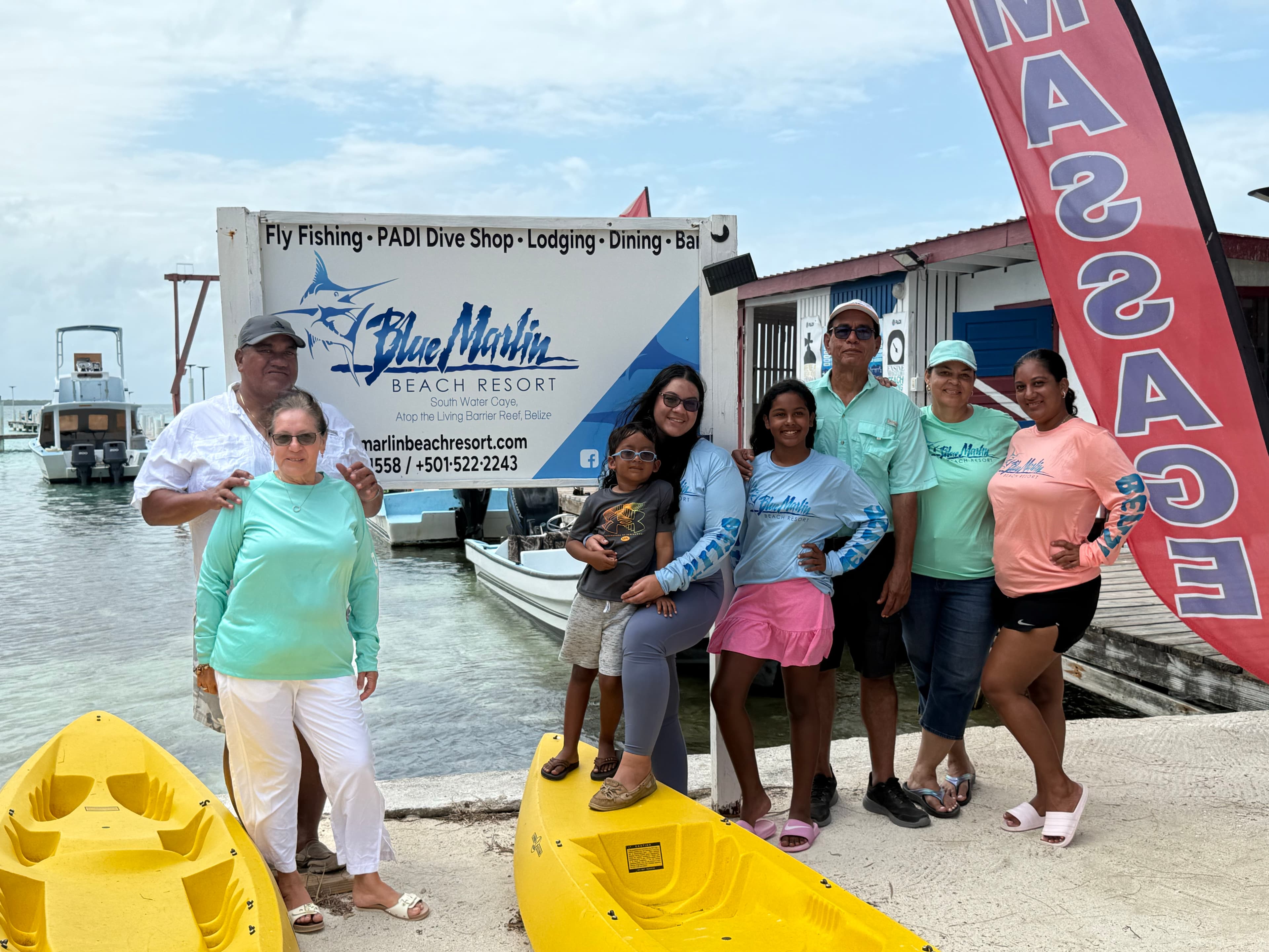 A diverse group of people stands in front of the Blue Marlin Beach Resort sign, with yellow kayaks in the foreground and boats in the background.