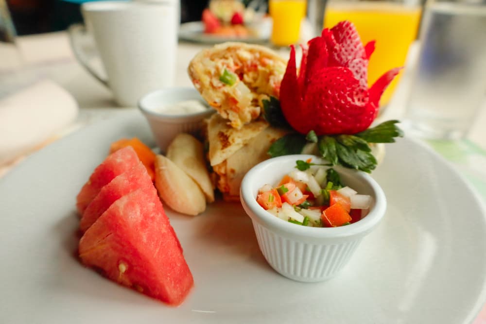 A plate with a rolled tortilla filled with colorful ingredients, accompanied by fresh fruit and a small bowl of salsa.