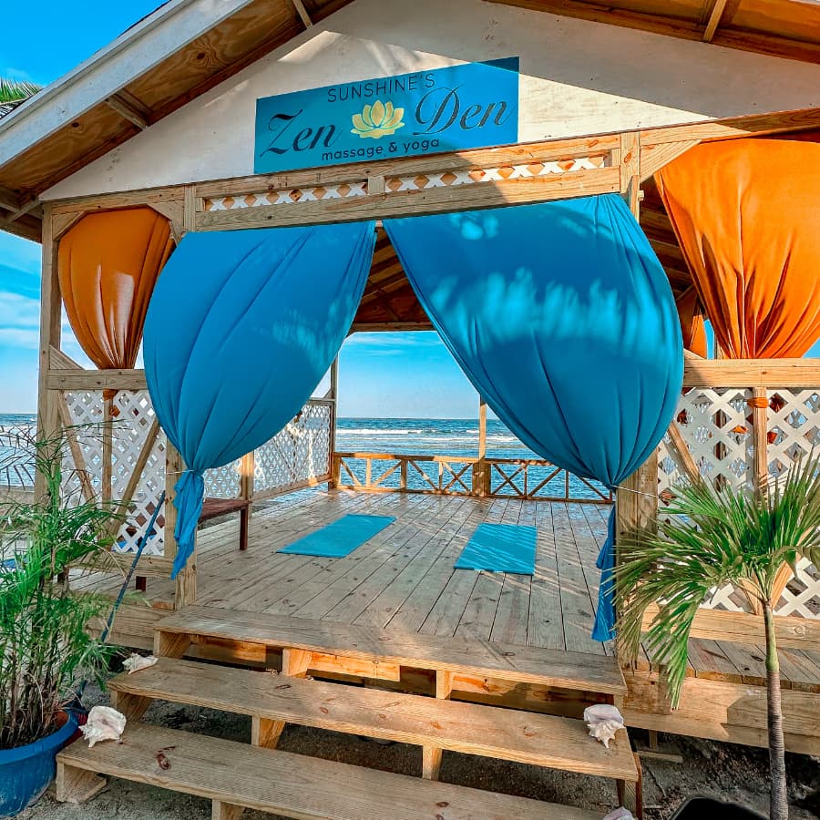 A wooden structure labeled "Sunshine's Zen Den massage & yoga" with vibrant blue and orange drapes, overlooking the ocean.