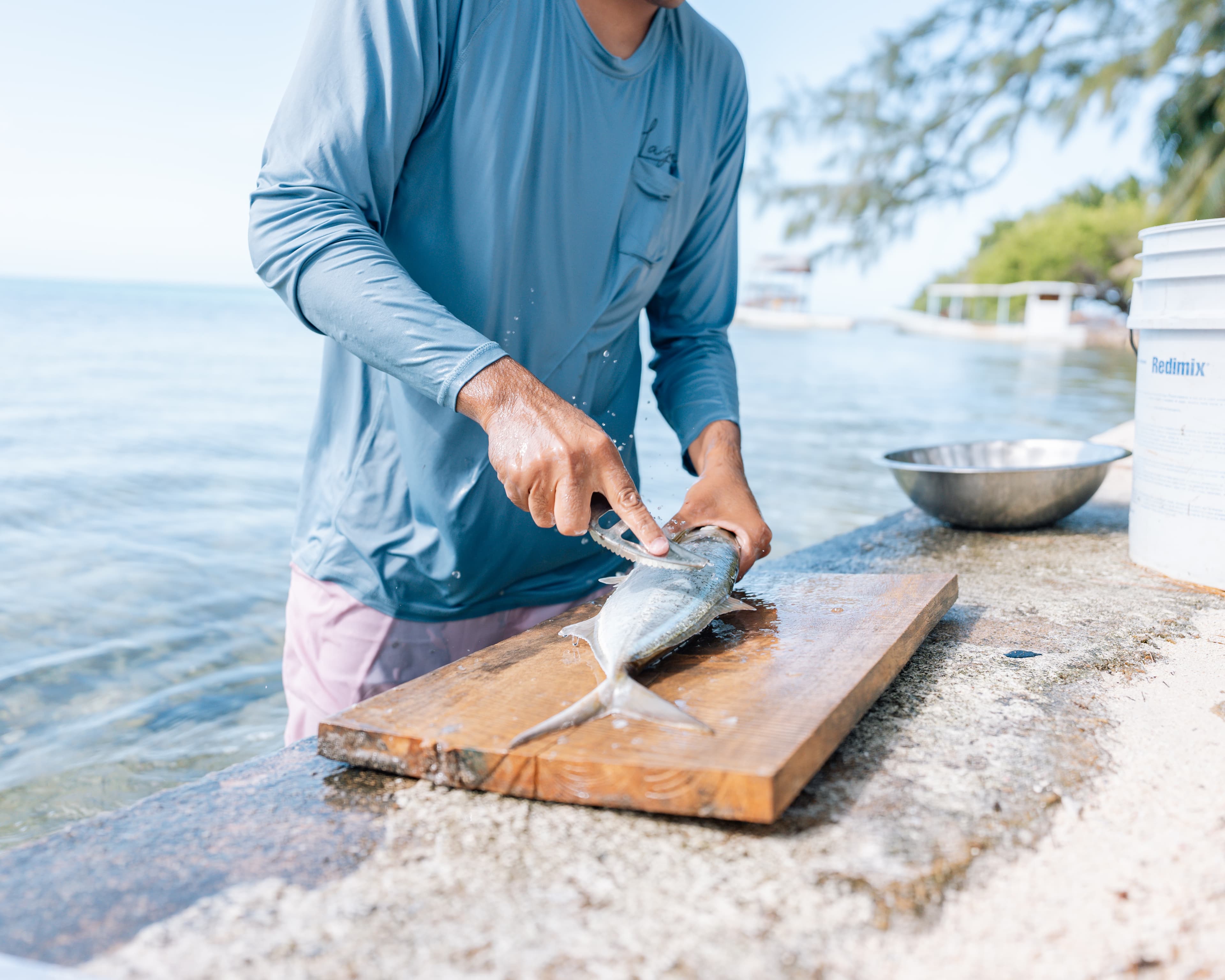 A person fillets a fish on a wooden cutting board by the water.