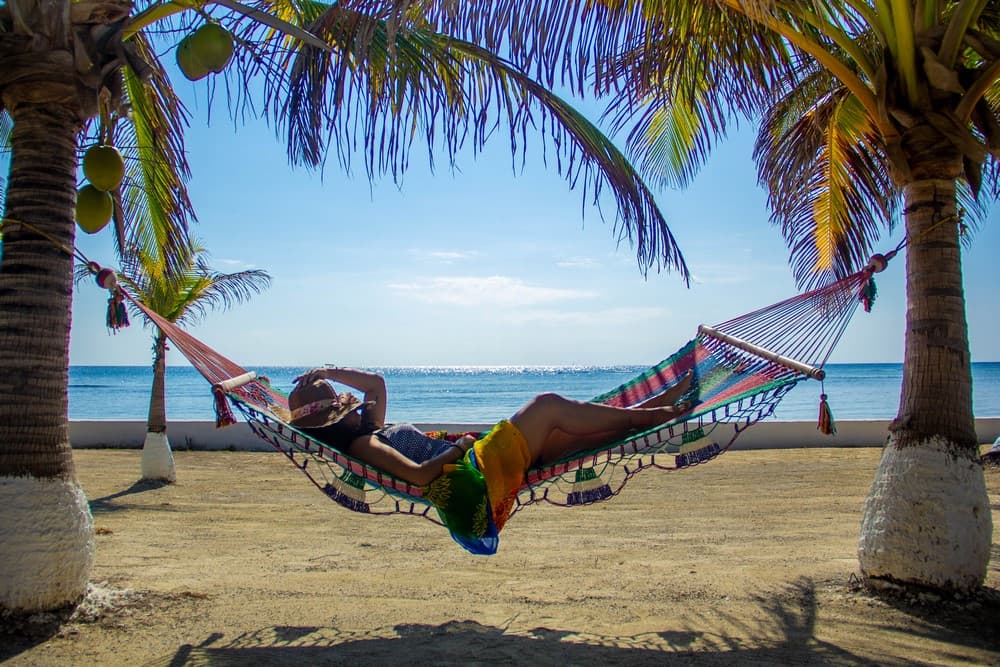 A person relaxes in a colorful hammock between palm trees on a beach by the ocean.