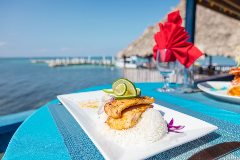 A plate of grilled fish garnished with lime sits on a turquoise table overlooking the ocean.