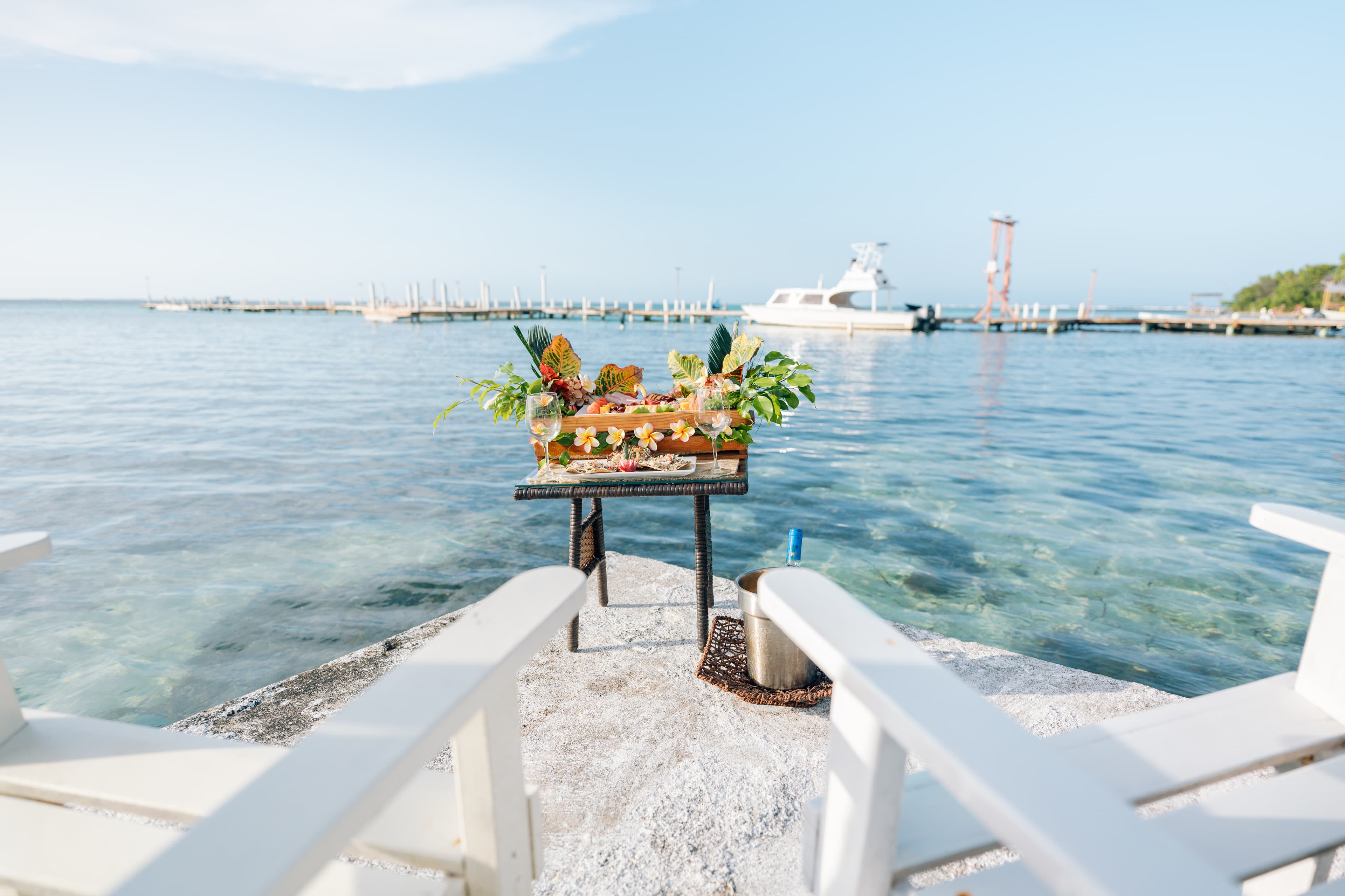 A table adorned with tropical fruits and garnishes overlooks clear water and a docked boat.
