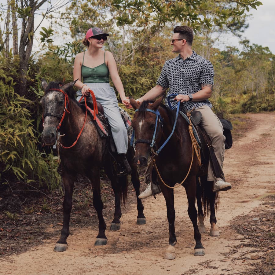 A couple shares a joyful moment while riding horses on a lush, wooded trail.