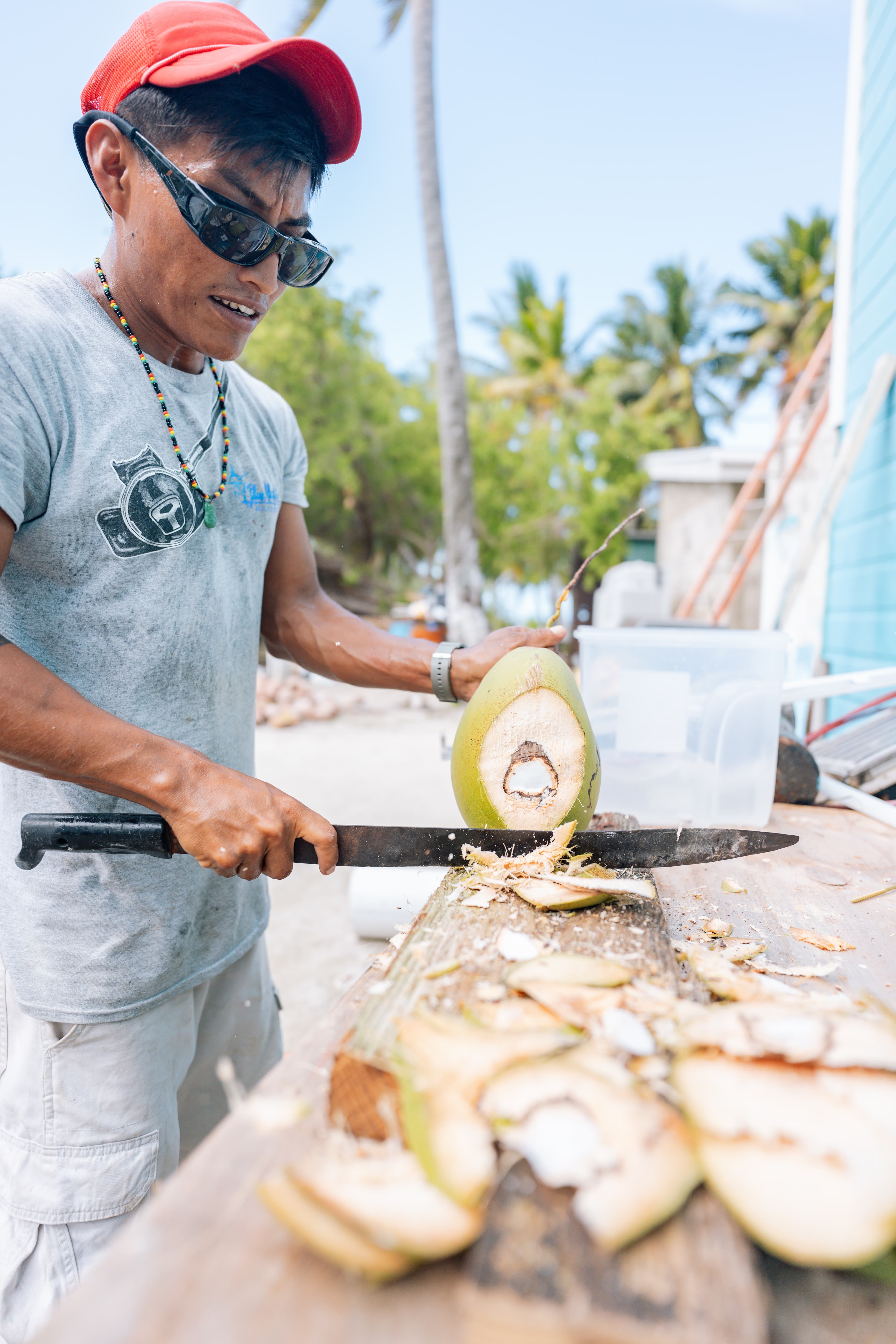 A man in a red cap cuts a fresh green coconut on a wooden table.