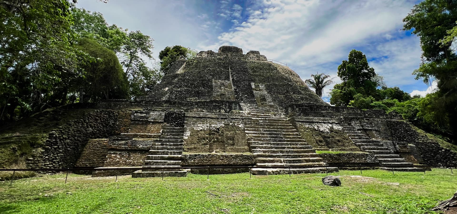Ancient stone pyramid surrounded by lush greenery and blue sky.