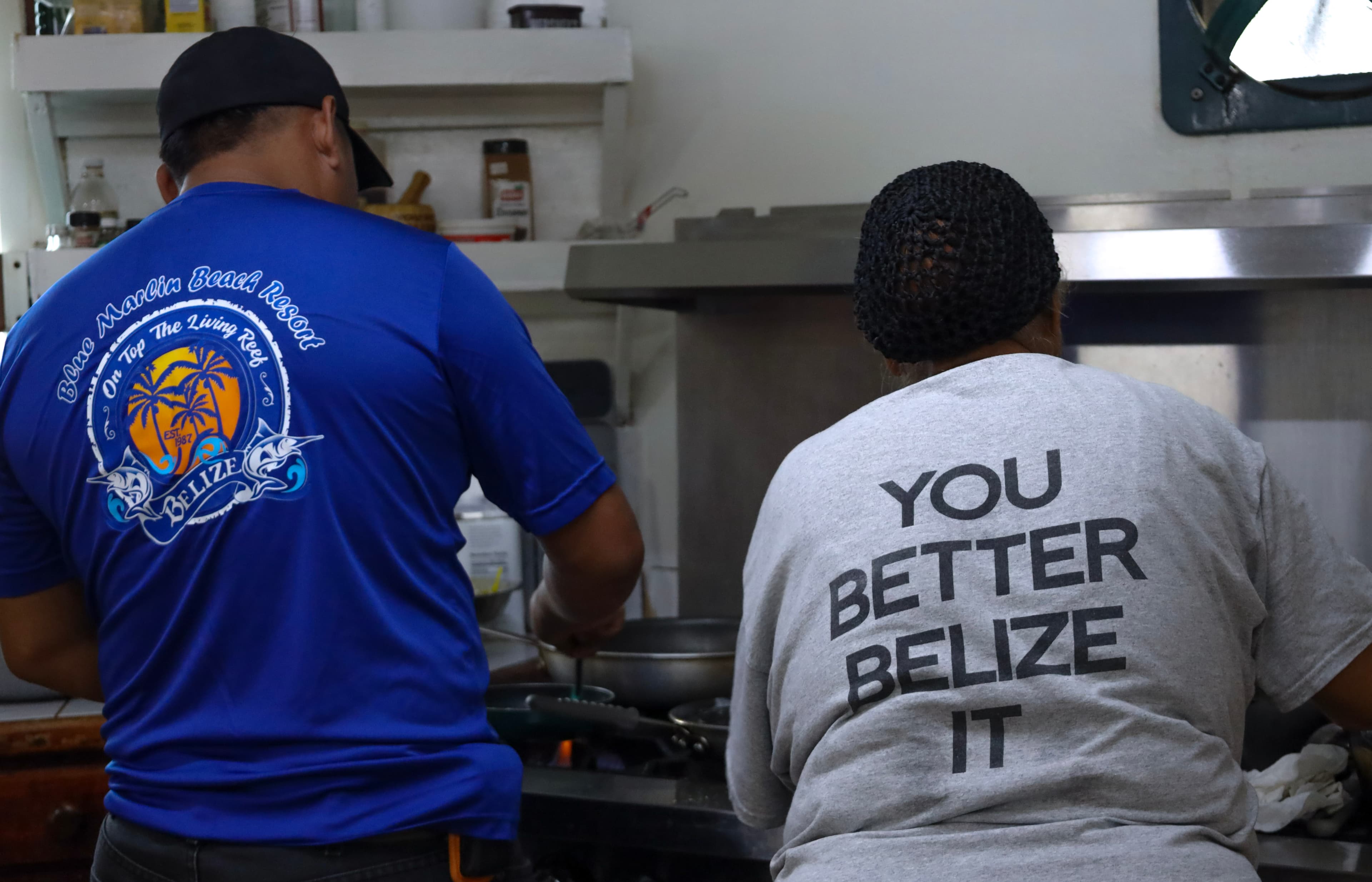 Two individuals cooking in a kitchen, one wearing a blue shirt and the other a gray shirt with "YOU BETTER BELIZE IT" printed on the back.