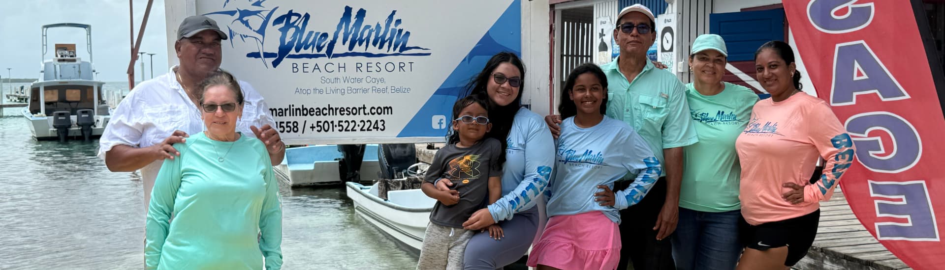 A group of people stands in front of a sign for Blue Marlin Beach Resort, with boats and water in the background.