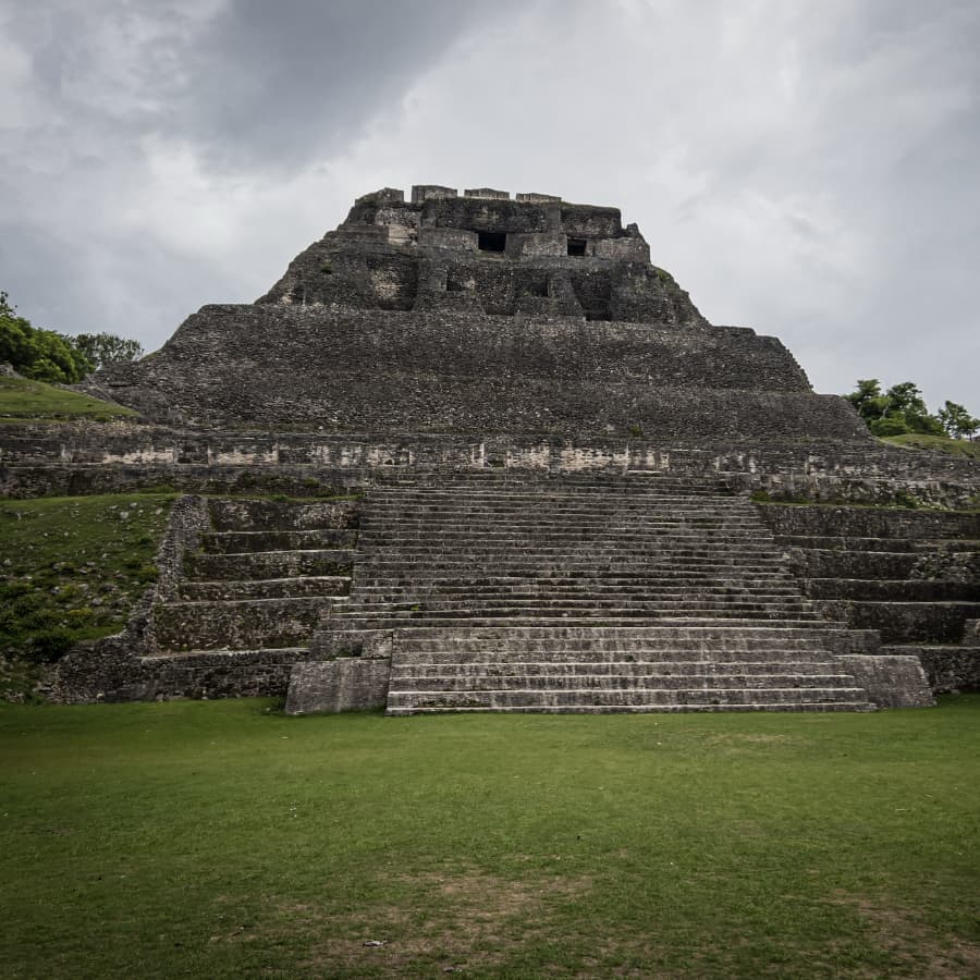 A stepped ancient structure rises from a grassy area under a cloudy sky.