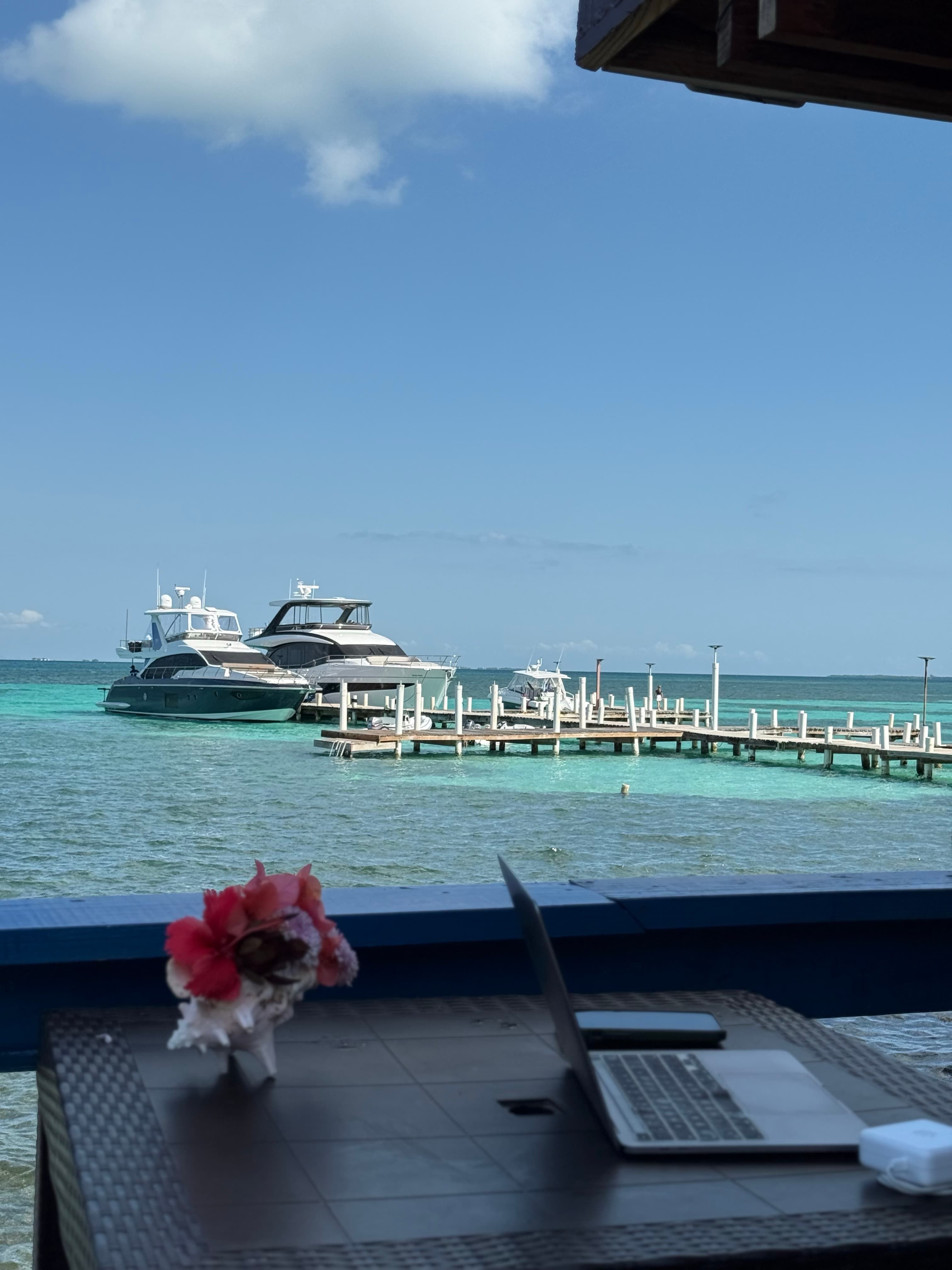 A laptop sits on a table with a floral centerpiece, overlooking boats at a dock on a sunny day.