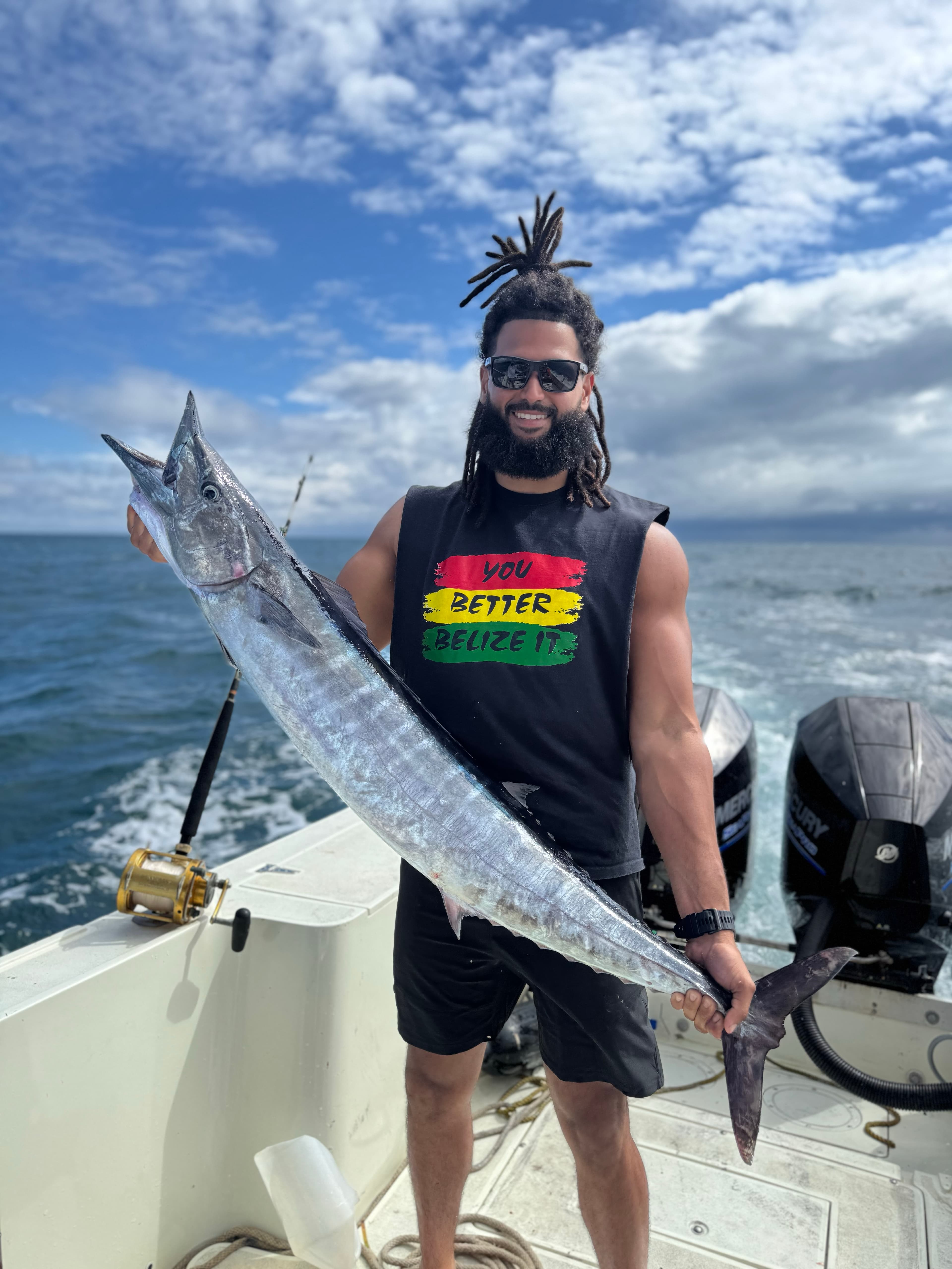 A man with dreadlocks smiles proudly while holding a large fish on a boat with a scenic ocean backdrop.