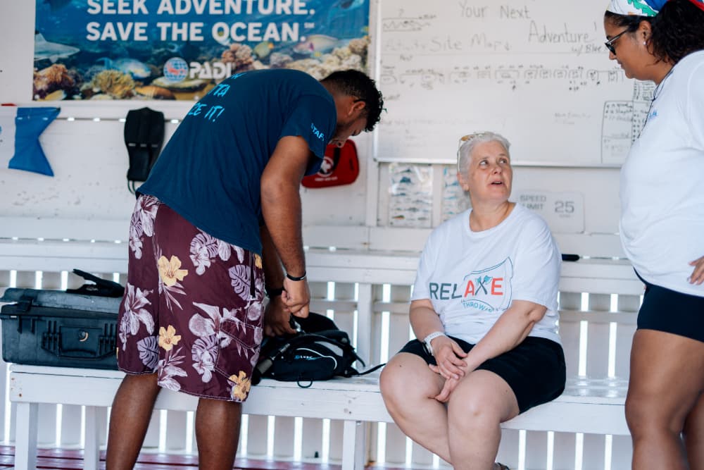 Three people are engaged in conversation in a diving center, with one preparing gear.