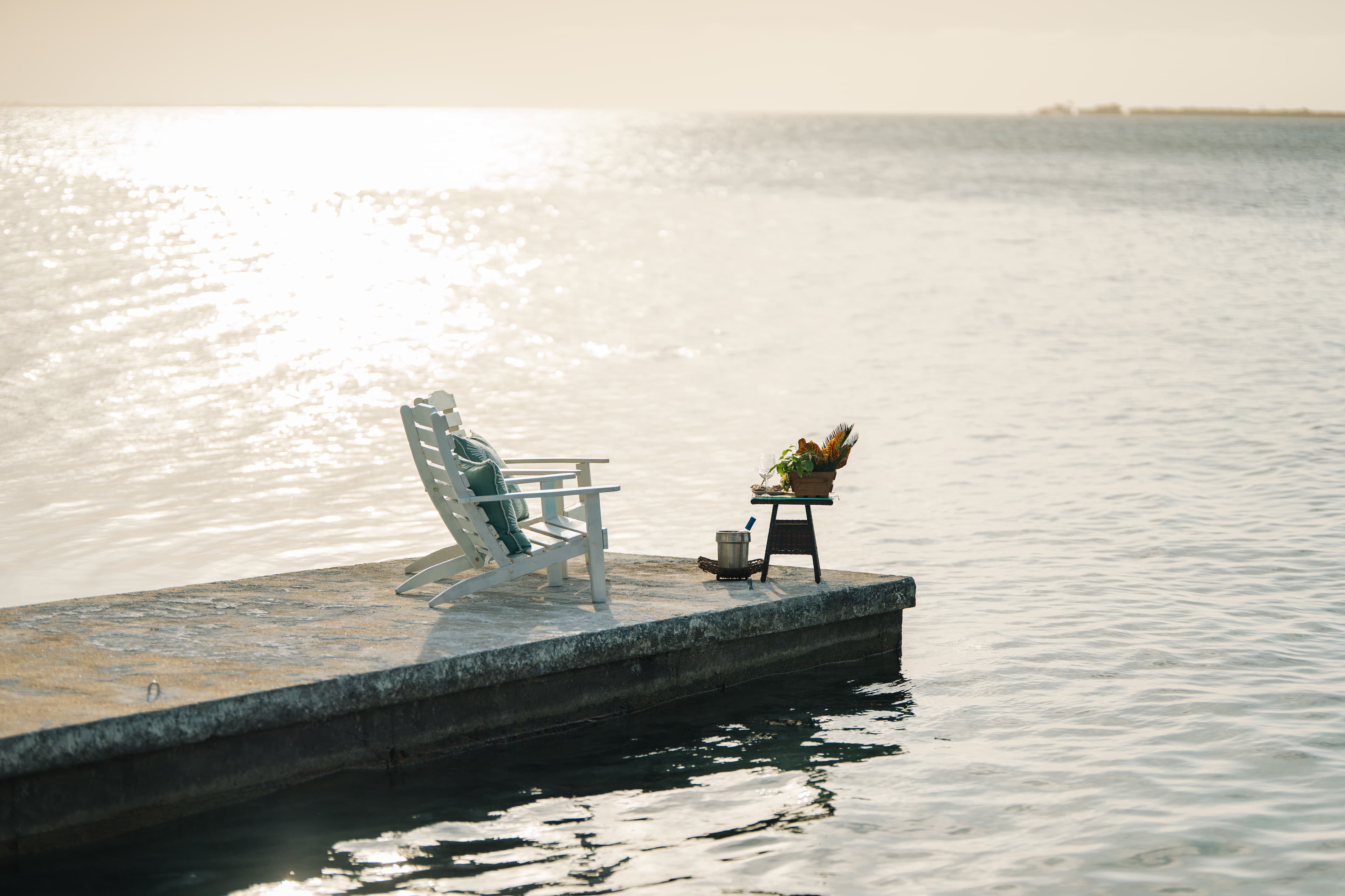 A rocking chair and a small table with plants sit on a tranquil dock overlooking the water.