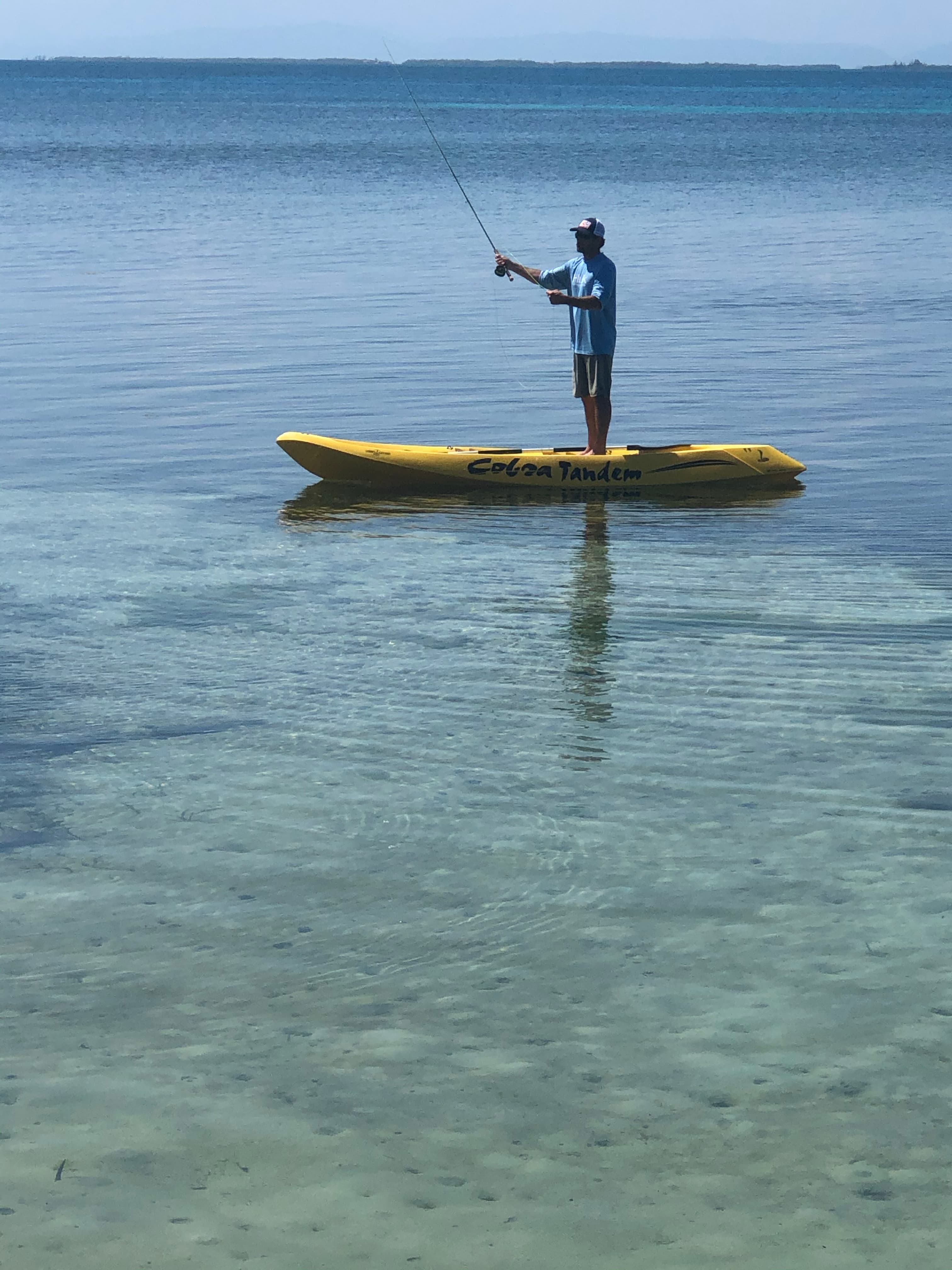 A person fishing from a yellow paddleboard in calm, clear waters.