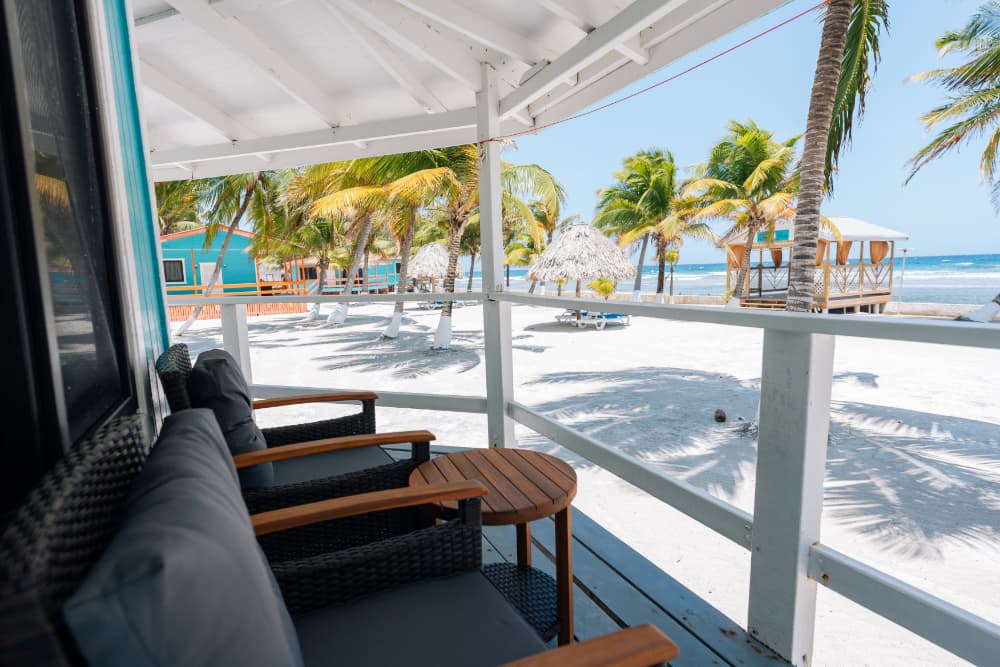 A cozy beachside porch with chairs facing a sandy shore and palm trees.