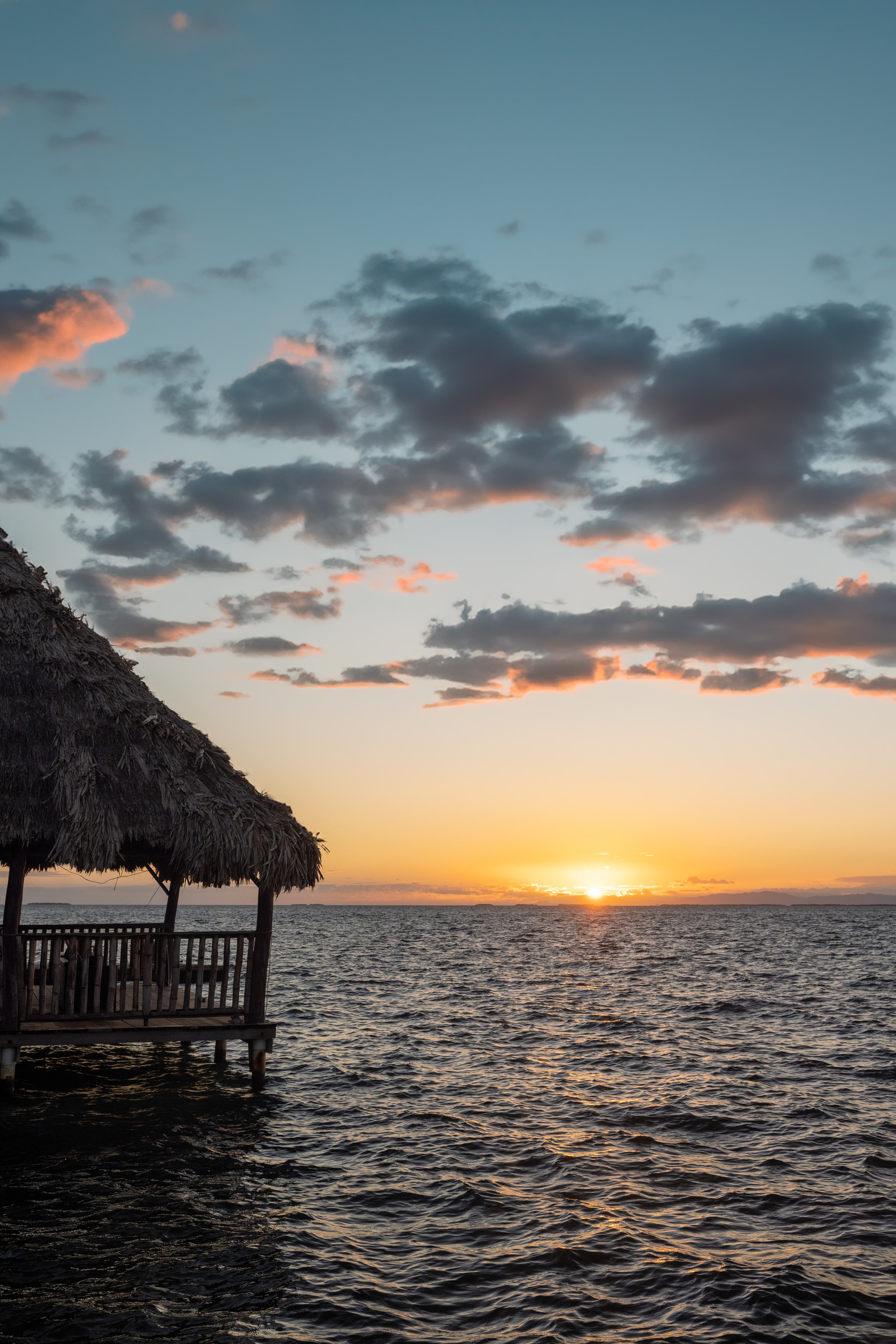 A thatched-roof hut overlooks the shimmering water at sunset, with colorful clouds in the sky.