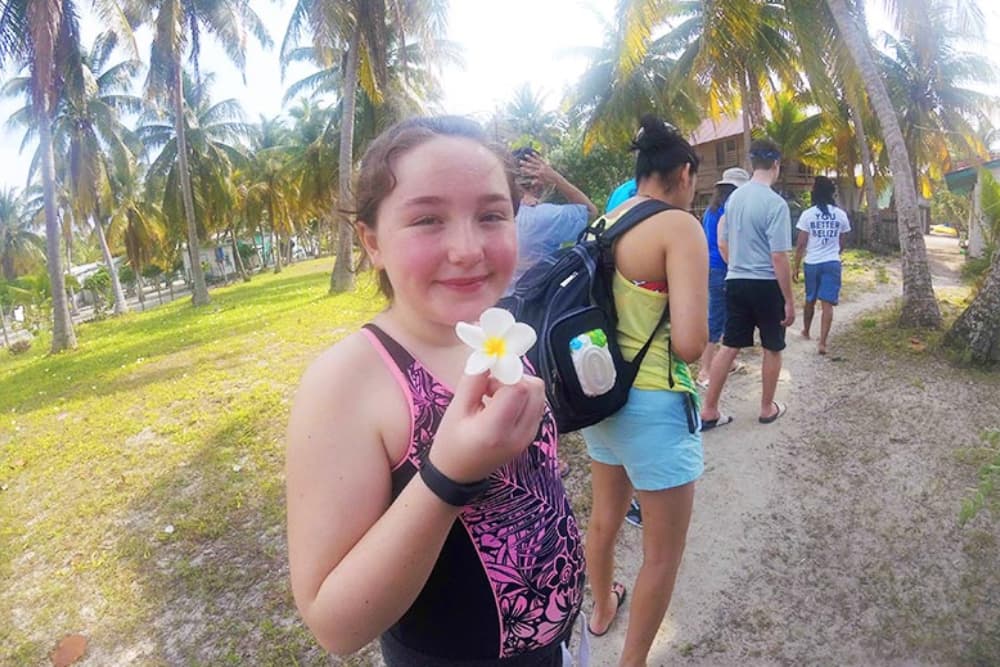 A smiling girl in a swimsuit holds a flower while walking along a palm tree-lined path with others.