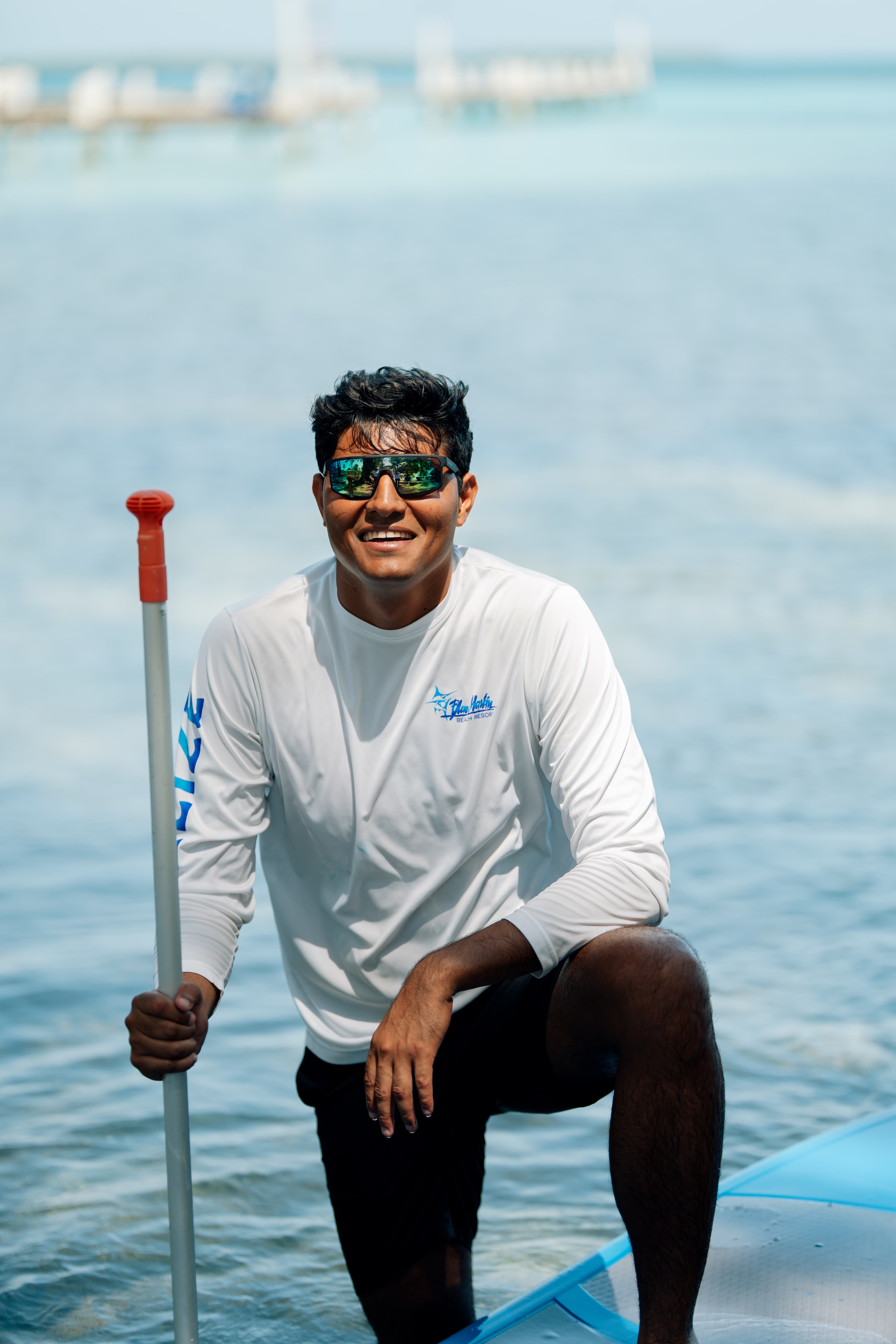 A man in a white long-sleeve shirt and sunglasses kneels on a paddleboard in calm water.