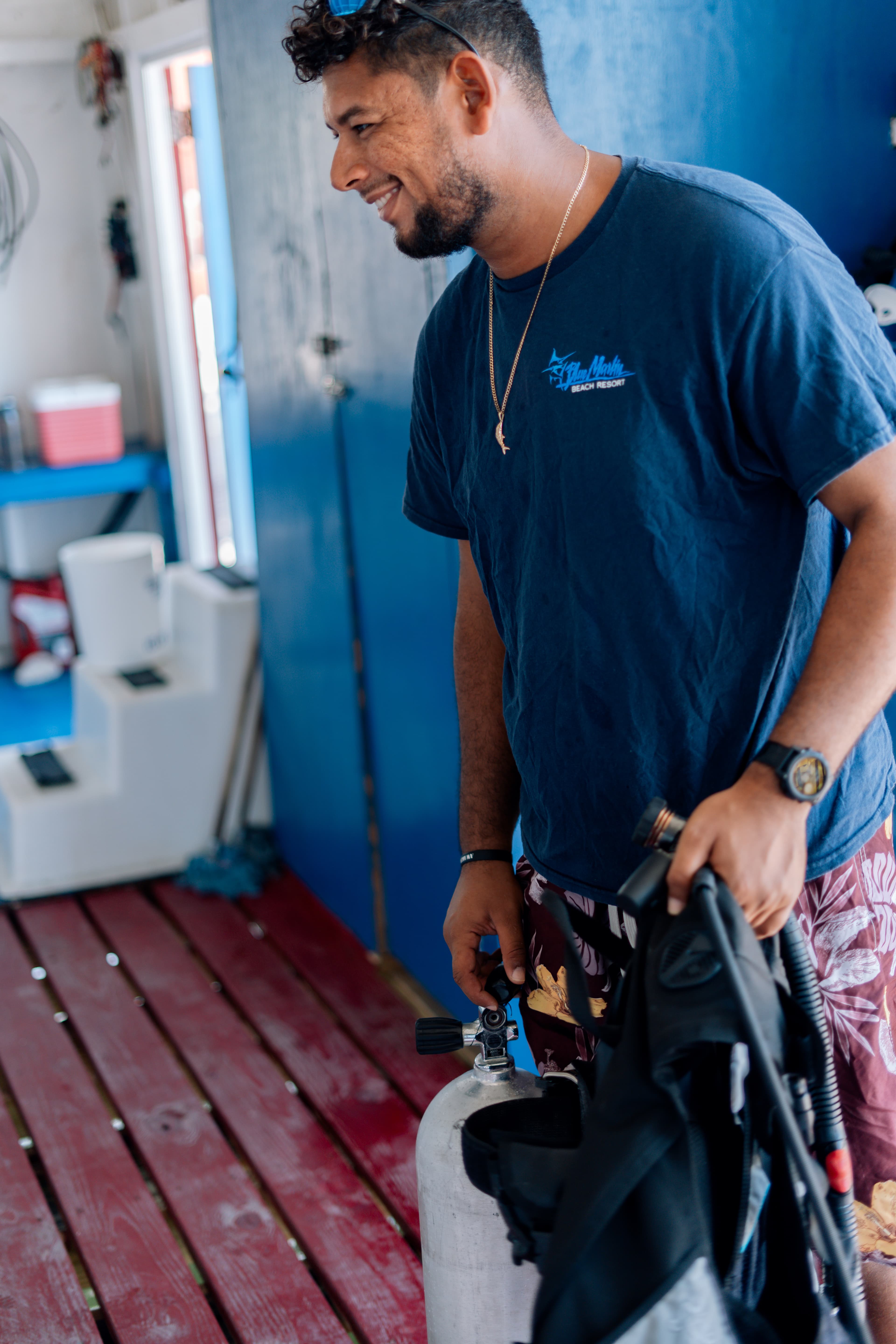 A smiling man in a blue shirt holds a scuba tank and diving gear inside a dive shop.