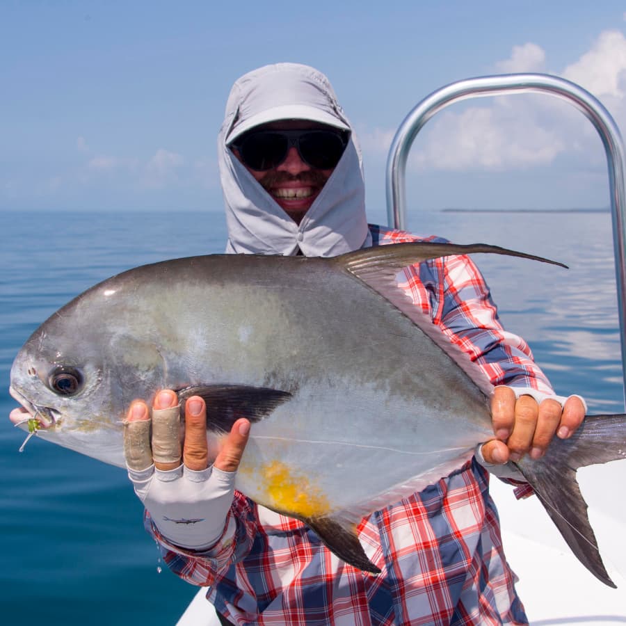 A smiling person in a sun hat and sunglasses holds a large fish on a boat with calm waters in the background.