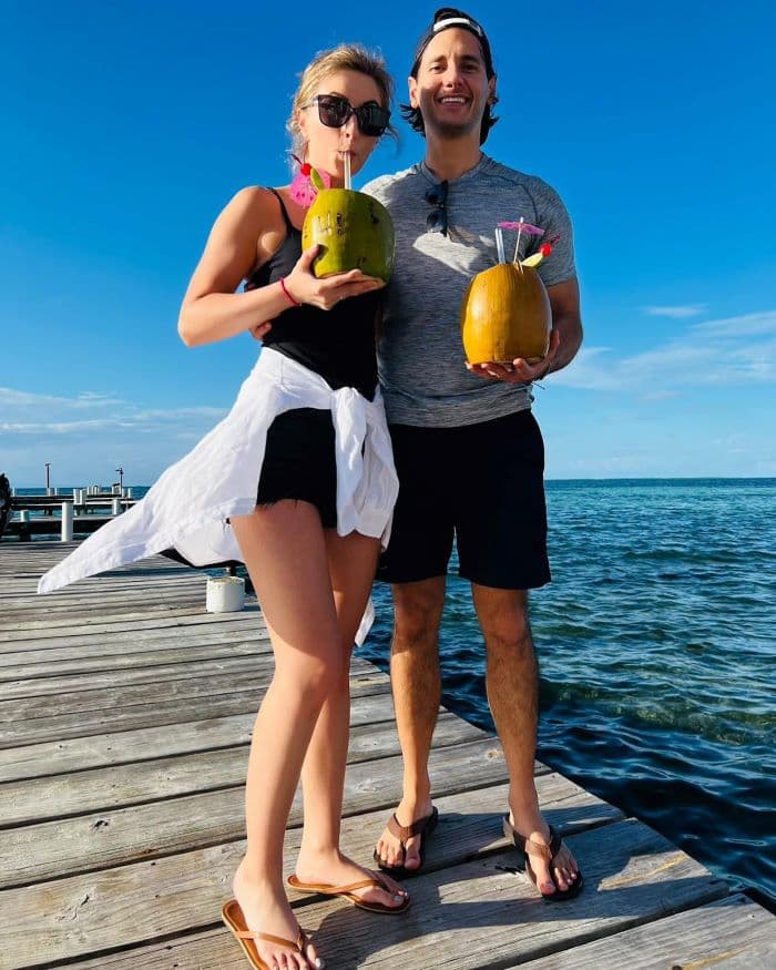 A couple stands on a dock holding coconuts with drinks, smiling against a sunny sky and ocean backdrop. A couple stands on a dock holding coconuts with drinks, smiling against a sunny sky and ocean backdrop.