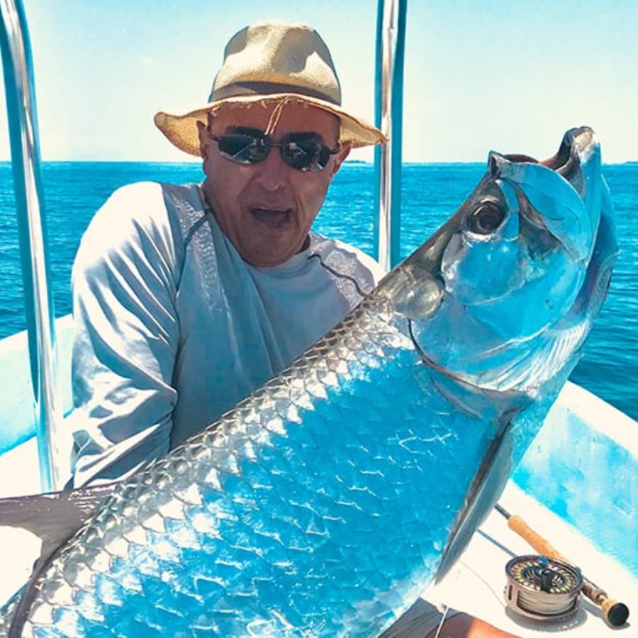 A man in a straw hat and sunglasses proudly holds a large fish while sitting in a boat on the water.