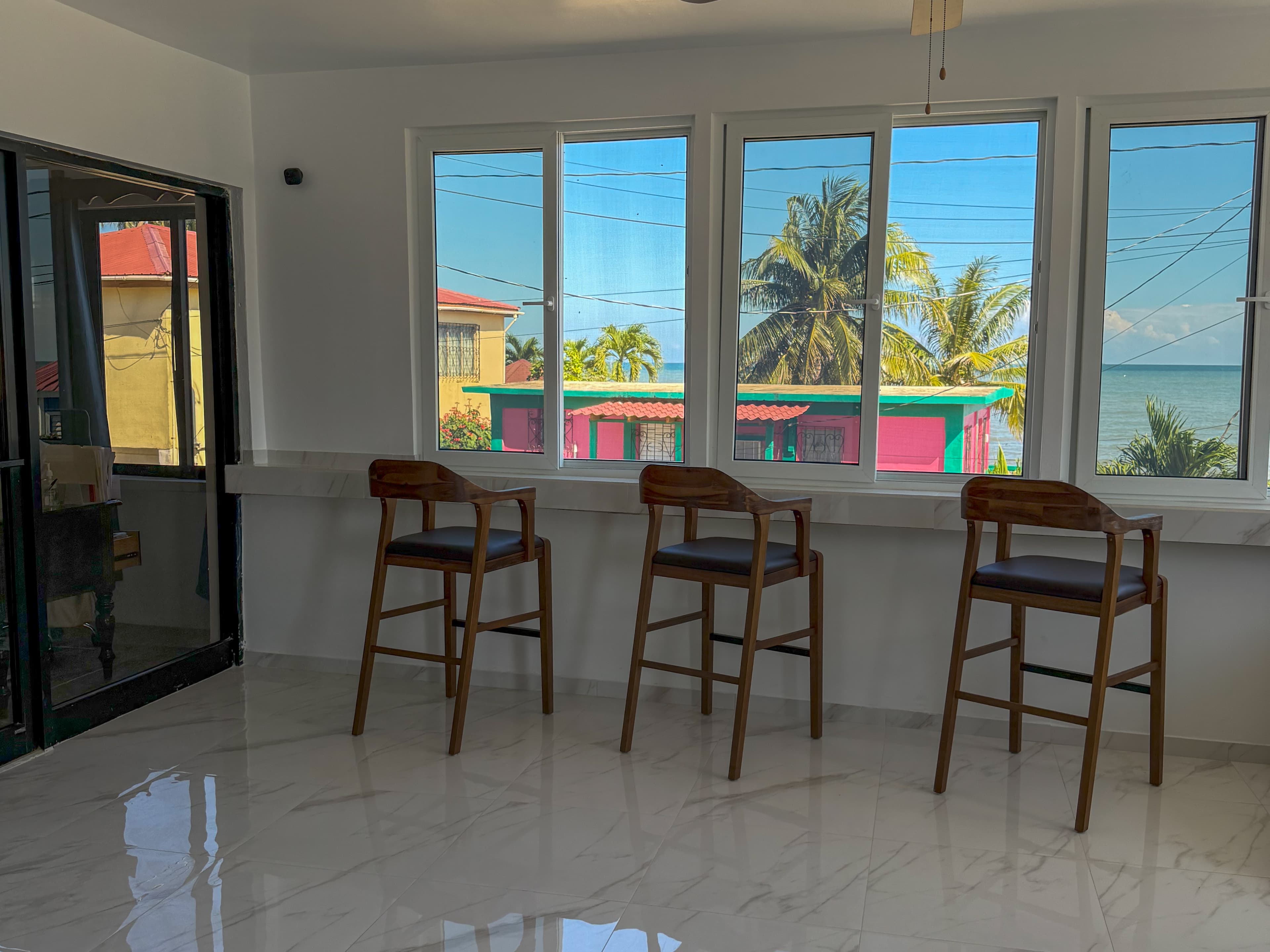 A bright room with three wooden bar stools facing large windows showcasing palm trees and colorful buildings by the sea.