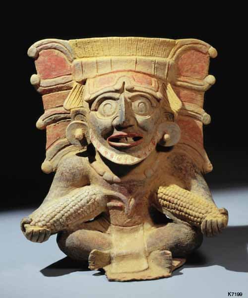 A colorful ancient ceramic figure with a grotesque face, holding cobs of corn.