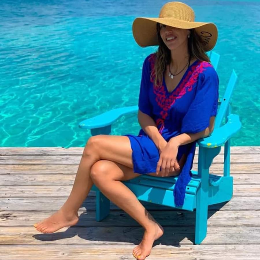 A woman in a blue dress and large sunhat sits on a turquoise chair by the water.