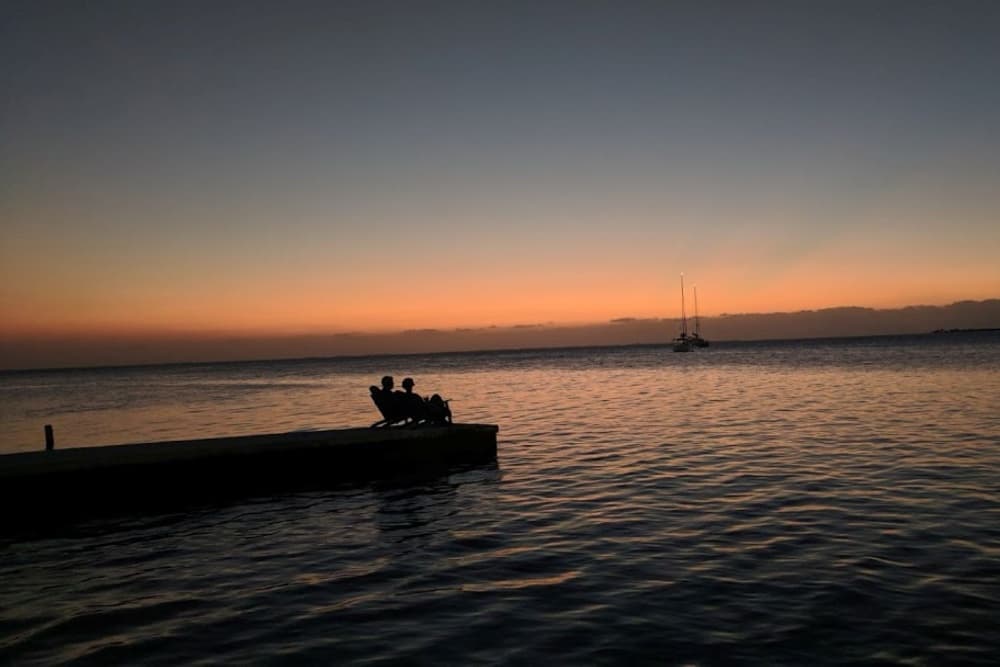 A couple sits on a dock watching a sunset over calm waters with a sailboat in the distance.