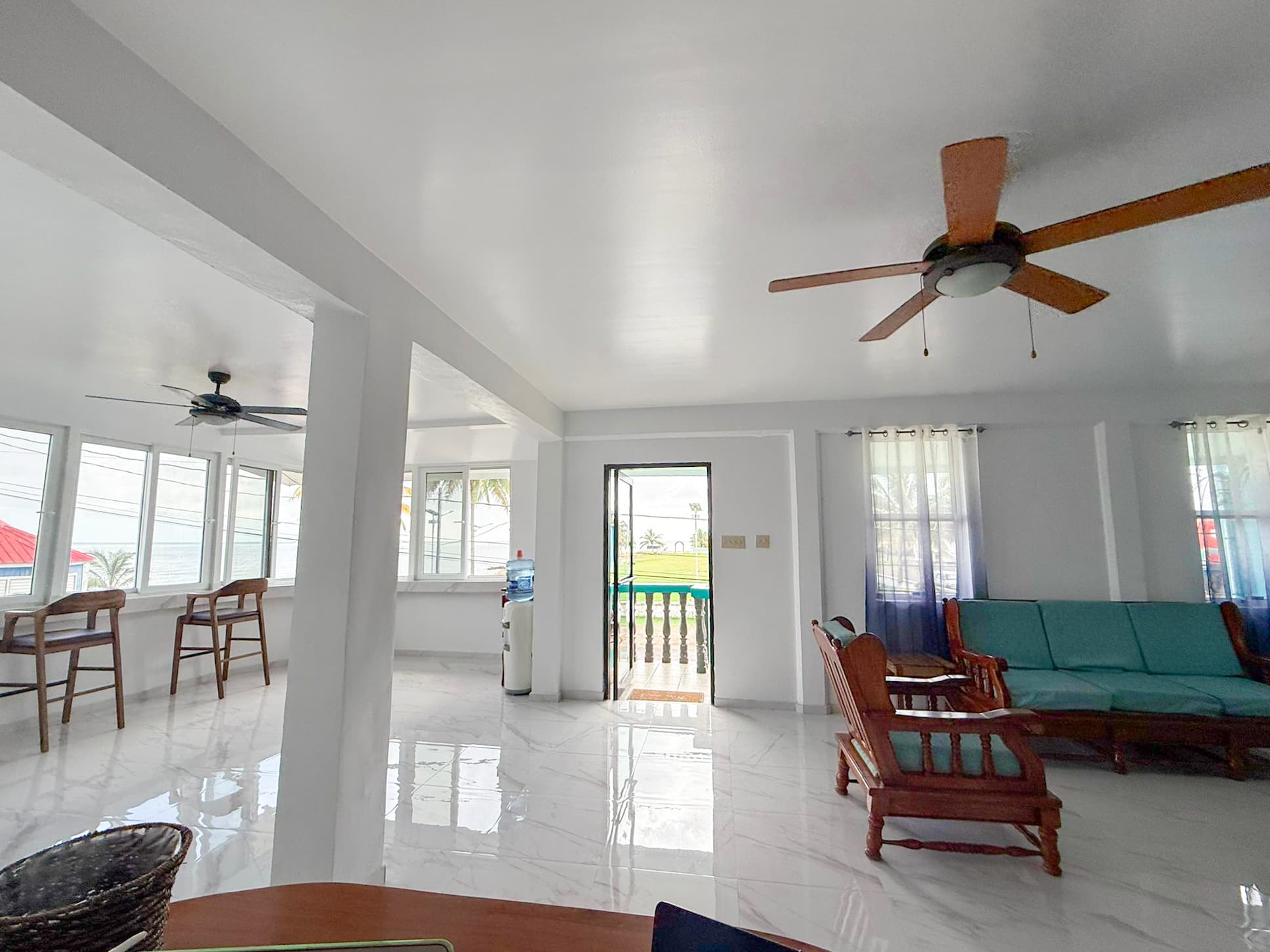 A bright, airy living room with white walls, tile flooring, and casual seating, overlooking outdoor greenery.