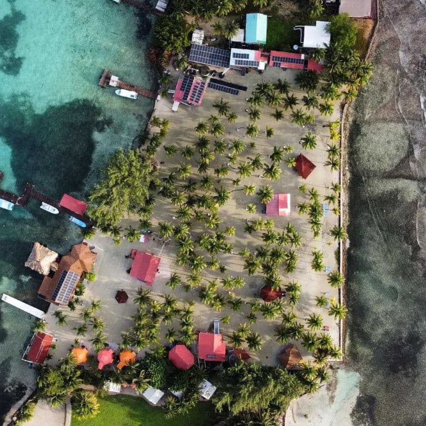 An aerial view of a private island with white sand beaches, dotted with palm trees and private cabanas, and the ocean surrounding the island.